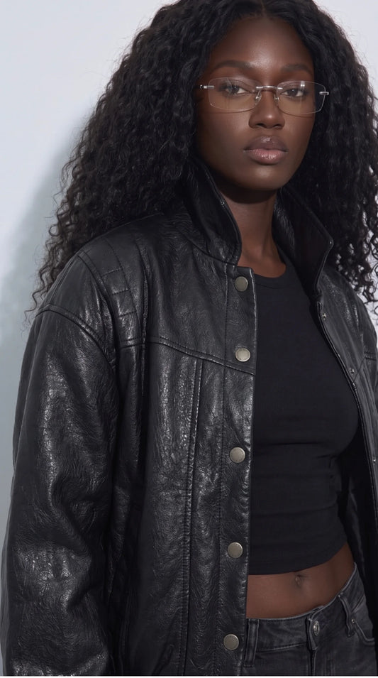 Detailed look at a black leather cropped jacket featuring quilted shoulder panels, curved seam lines, a structured collar and a row of brushed-metal snap fasteners. The naturally textured leather catches the light across the sleeves and body, creating a distinctive blend of modern moto-inspired design and understated luxury for women’s statement outerwear.