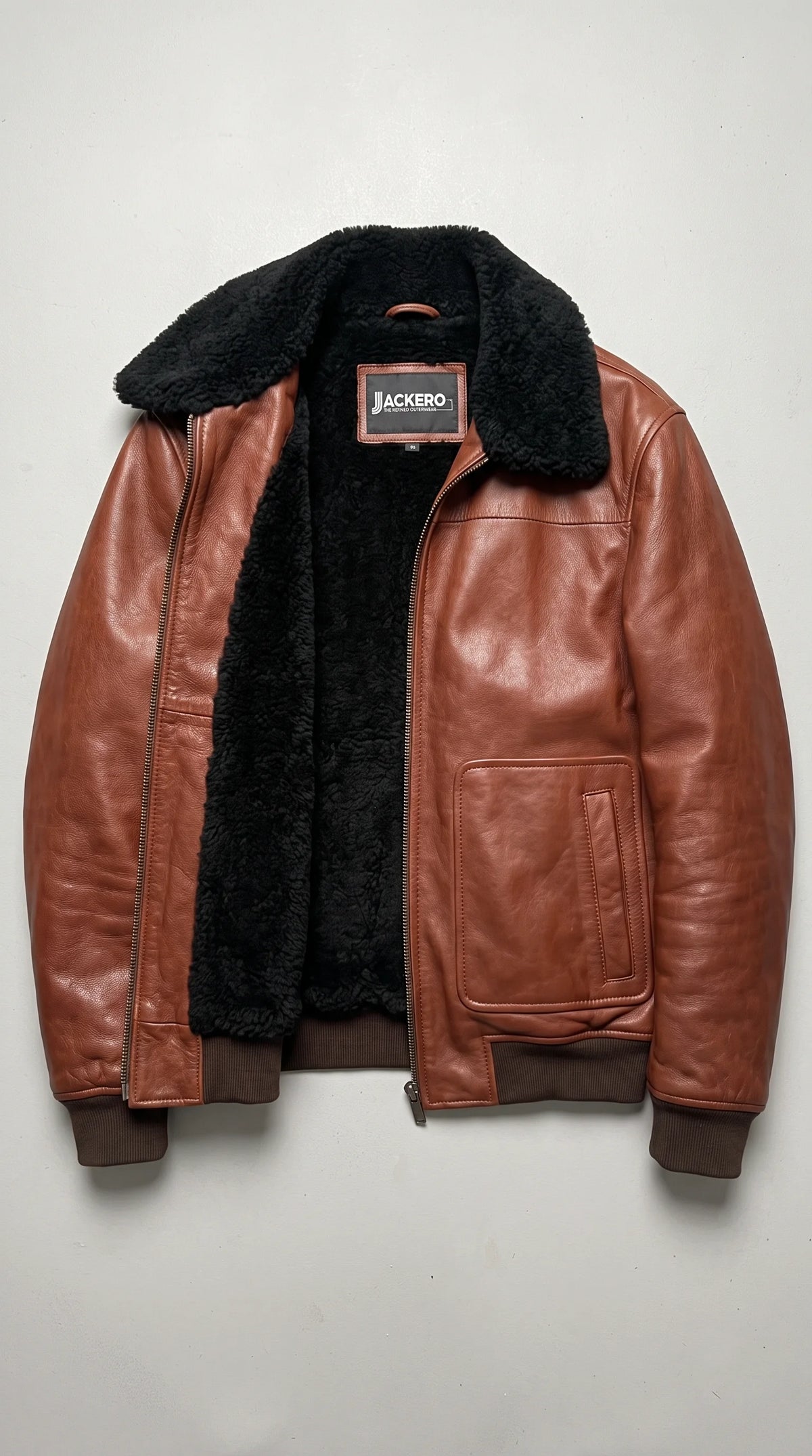 Cognac-brown genuine leather aviator jacket displayed open to reveal its black plush-look interior and JACKERO neck label, with a contrasting black shearling collar, full-length metal zipper, large lower patch pockets and dark ribbed trims.