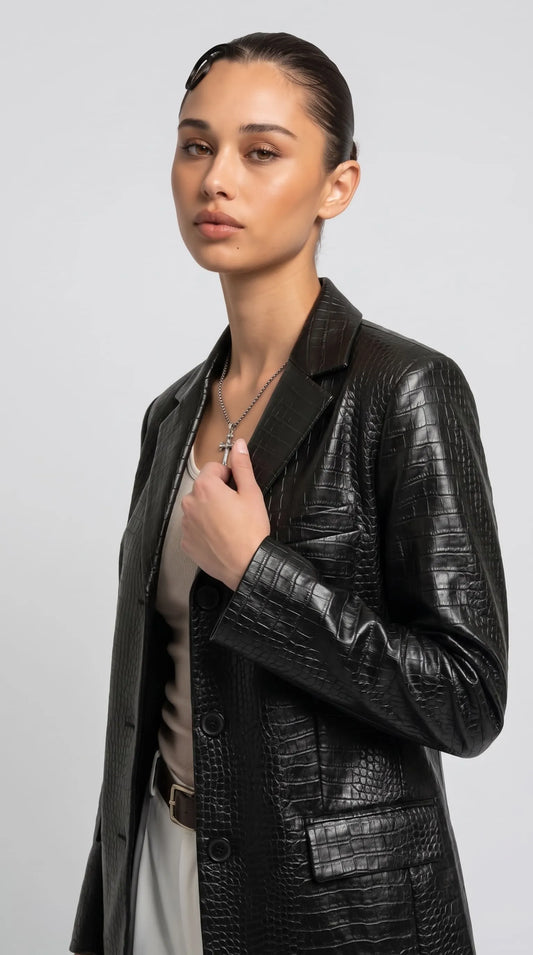 Close-up of a women's black crocodile-embossed leather blazer highlighting its raised reptile-inspired grain, wide notch lapels, dark front buttons and clean chest welt pocket. Reflective highlights across the textured leather emphasise its bold fashion character, while the tailored construction and refined detailing make this statement blazer suitable for polished monochrome outfits and contemporary designer-inspired styling.
