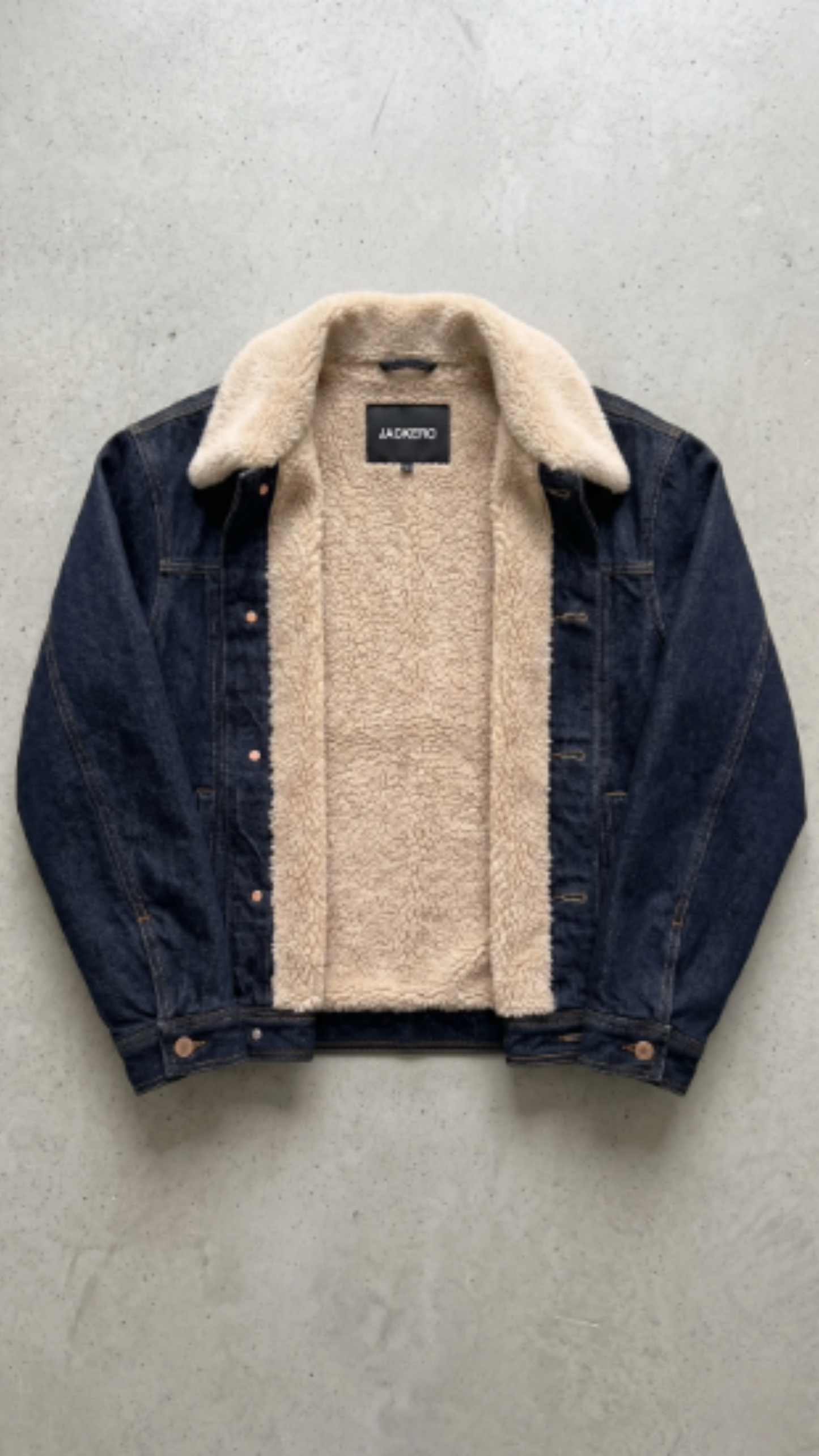 A dark blue denim jacket with a thick beige sherpa fleece lining, laid flat on a textured gray concrete surface. The brand name 'JACKERO' is visible on the inner neck label.
