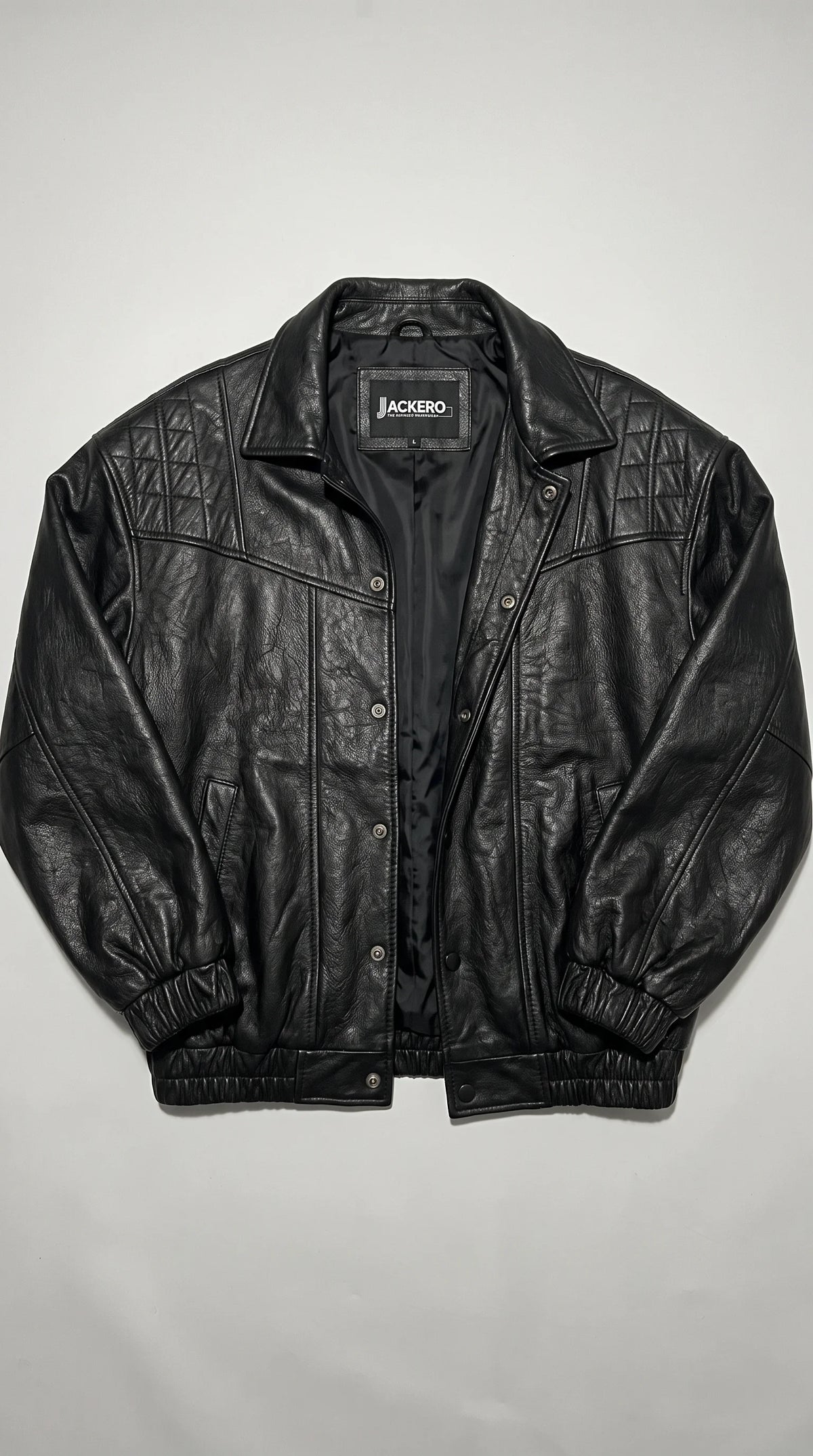 JACKERO women’s black genuine leather cropped jacket displayed open to show its smooth black inner lining, branded neck label, snap-button front and quilted shoulder panels. The structured collar, vertical stitching and elasticated cuffs and hem complete its modern silhouette, combining distinctive leather texture with a clean, versatile finish for contemporary statement outerwear.