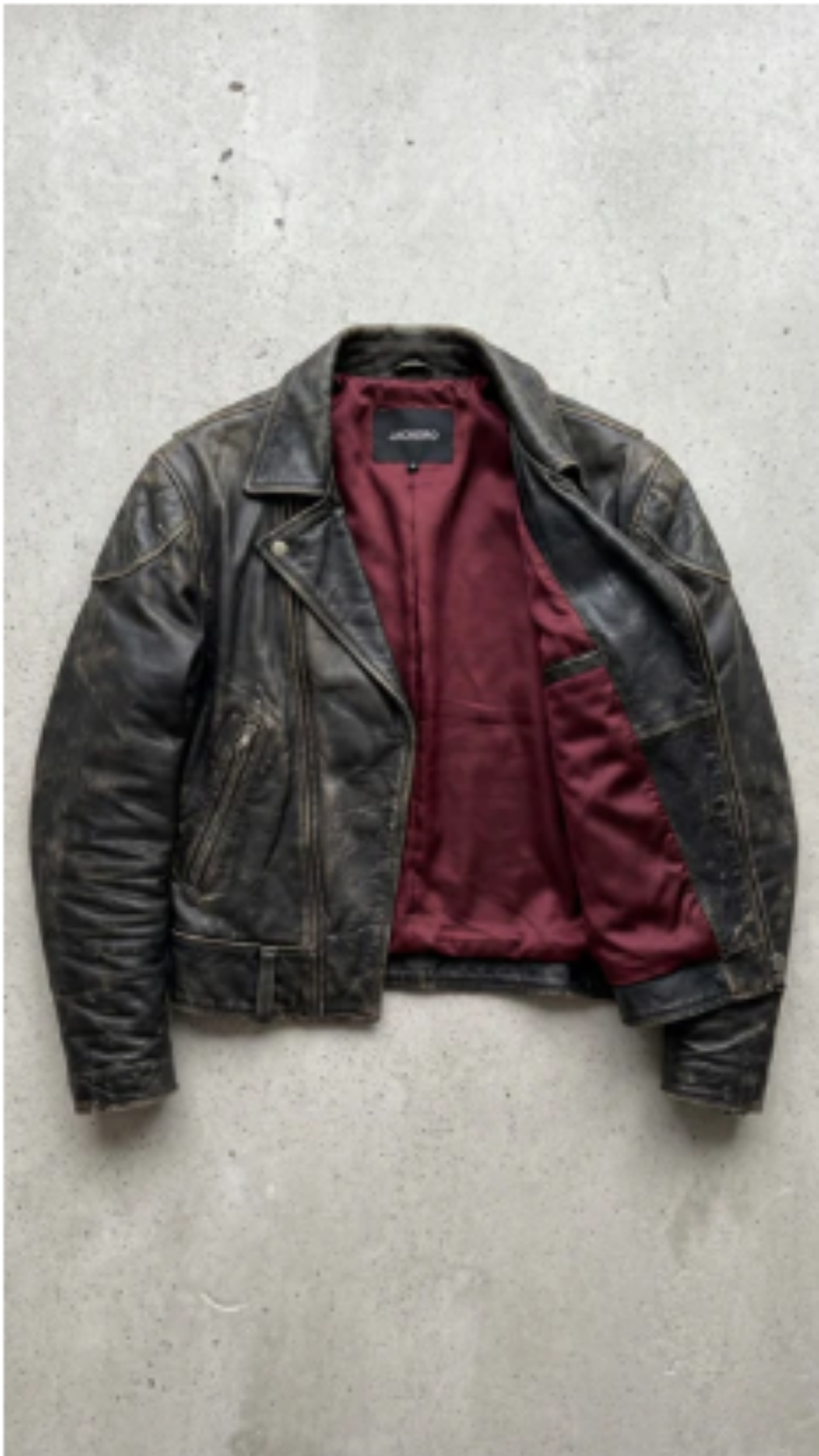 A distressed black leather motorcycle jacket with a deep burgundy red lining, laid flat on a textured gray concrete surface.