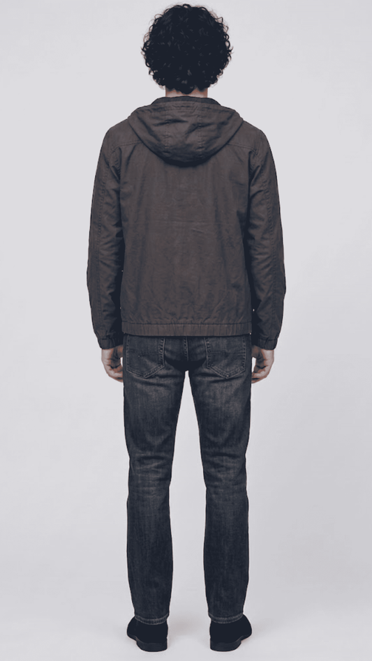 A man viewed from behind wearing a dark gray hooded jacket and blue jeans, standing against a light gray studio background.