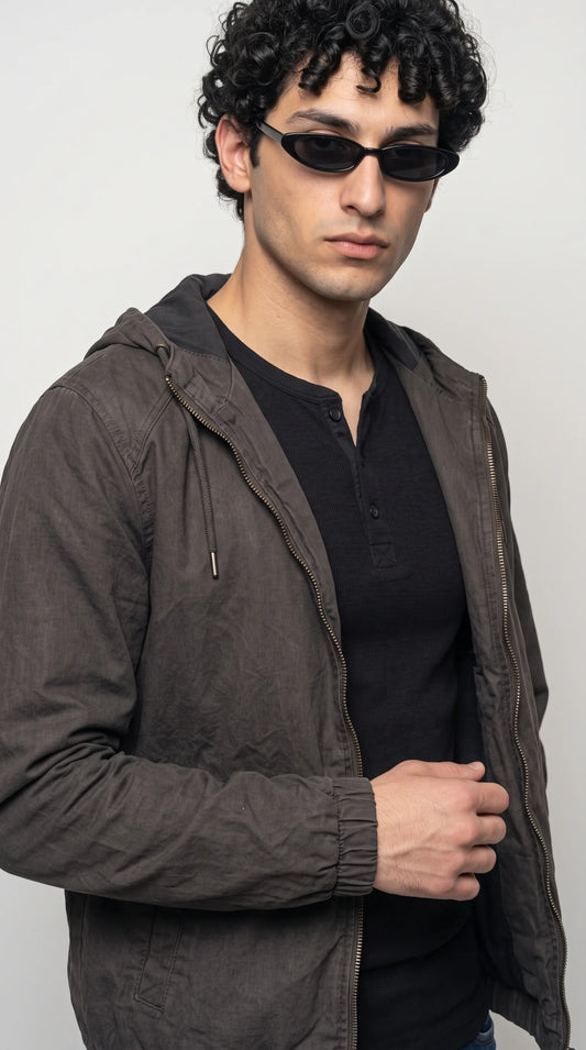 Smoky dark-grey hooded jacket featuring an adjustable drawstring hood, metal-tipped cords, a full-length front zipper and subtly textured silk-cotton fabric. Its muted charcoal tone and clean construction create a refined utility-inspired finish, making this lightweight men’s zip-up jacket an easy choice for minimalist casual outfits and contemporary off-duty styling.