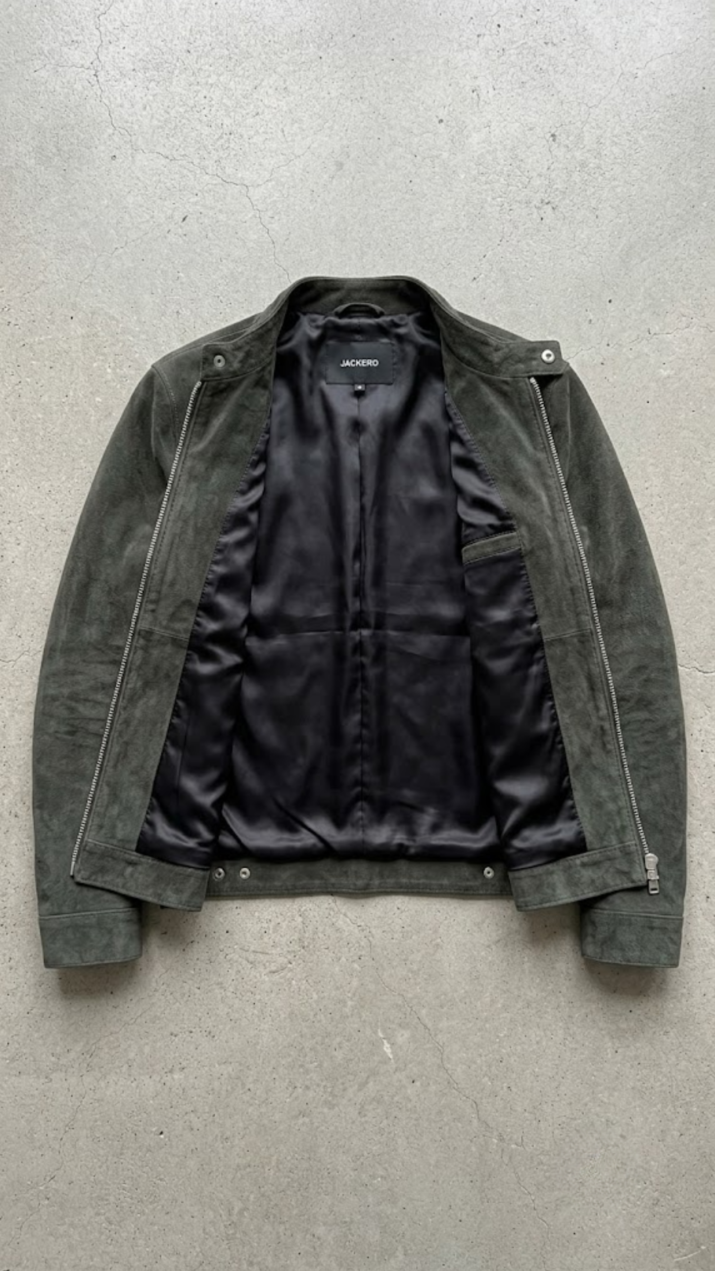 A dark olive green suede jacket with a black satin lining, laid flat on a textured gray concrete surface.