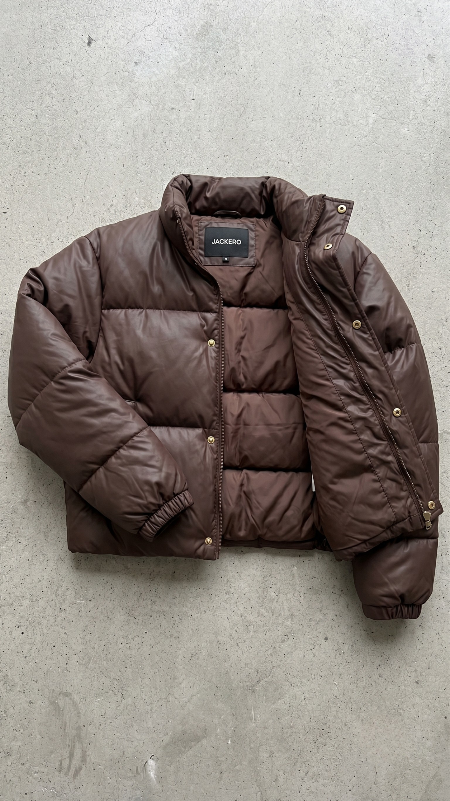 Close-up shot of a dark chocolate brown puffer jacket showing the premium textured fabric, zipper closure, and padded collar.