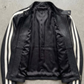 A black leather jacket with white stripes down the sleeves, laid flat on a gray concrete surface.