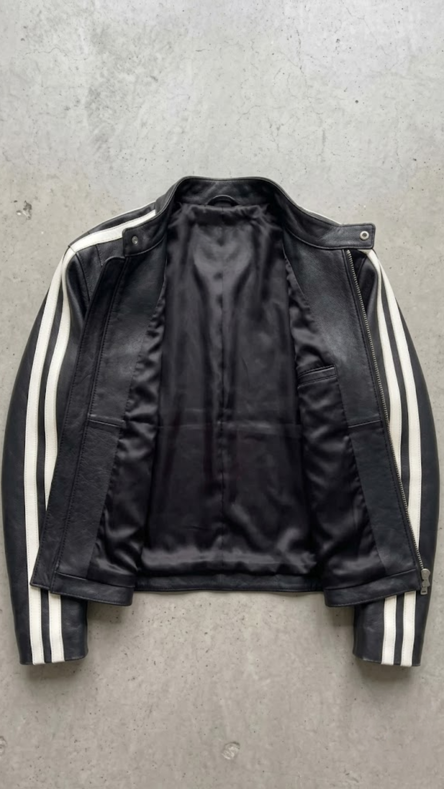 A black leather jacket with white stripes down the sleeves, laid flat on a gray concrete surface.