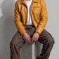 A model sits cross-legged on a concrete floor wearing a burgundy leather moto jacket, black jeans, and white sneakers against a neutral gray background.