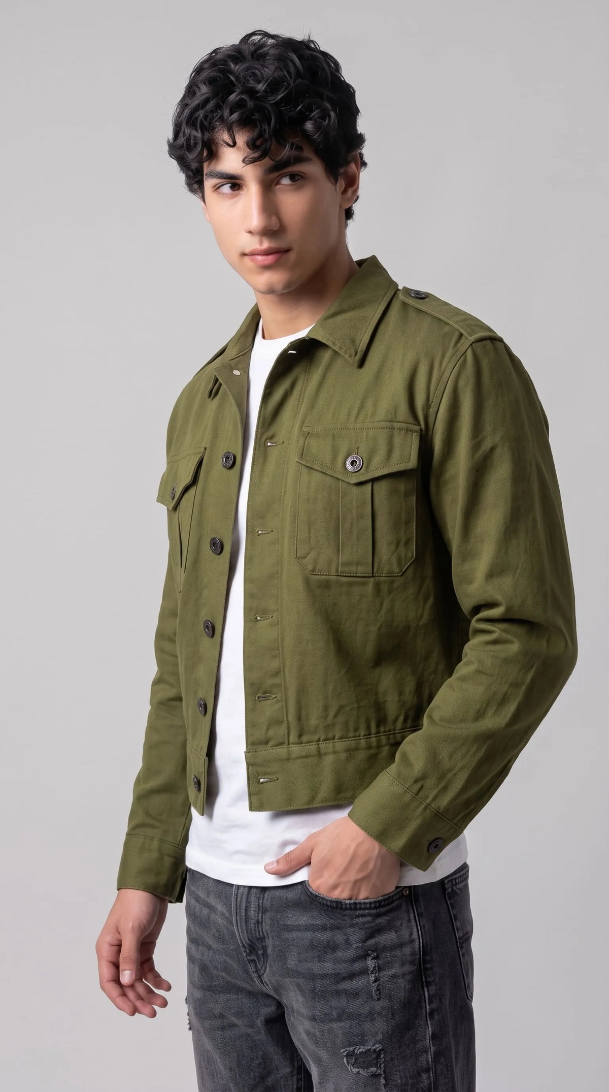 Army olive green heavyweight cotton canvas Ike jacket with buttoned military shoulder epaulettes, twin pleated flap chest pockets and contrasting black buttons, styled with a white T-shirt and charcoal jeans for modern heritage streetwear.