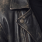 Close-up of a distressed black leather jacket with a pointed collar and silver snap button, worn over a black top.