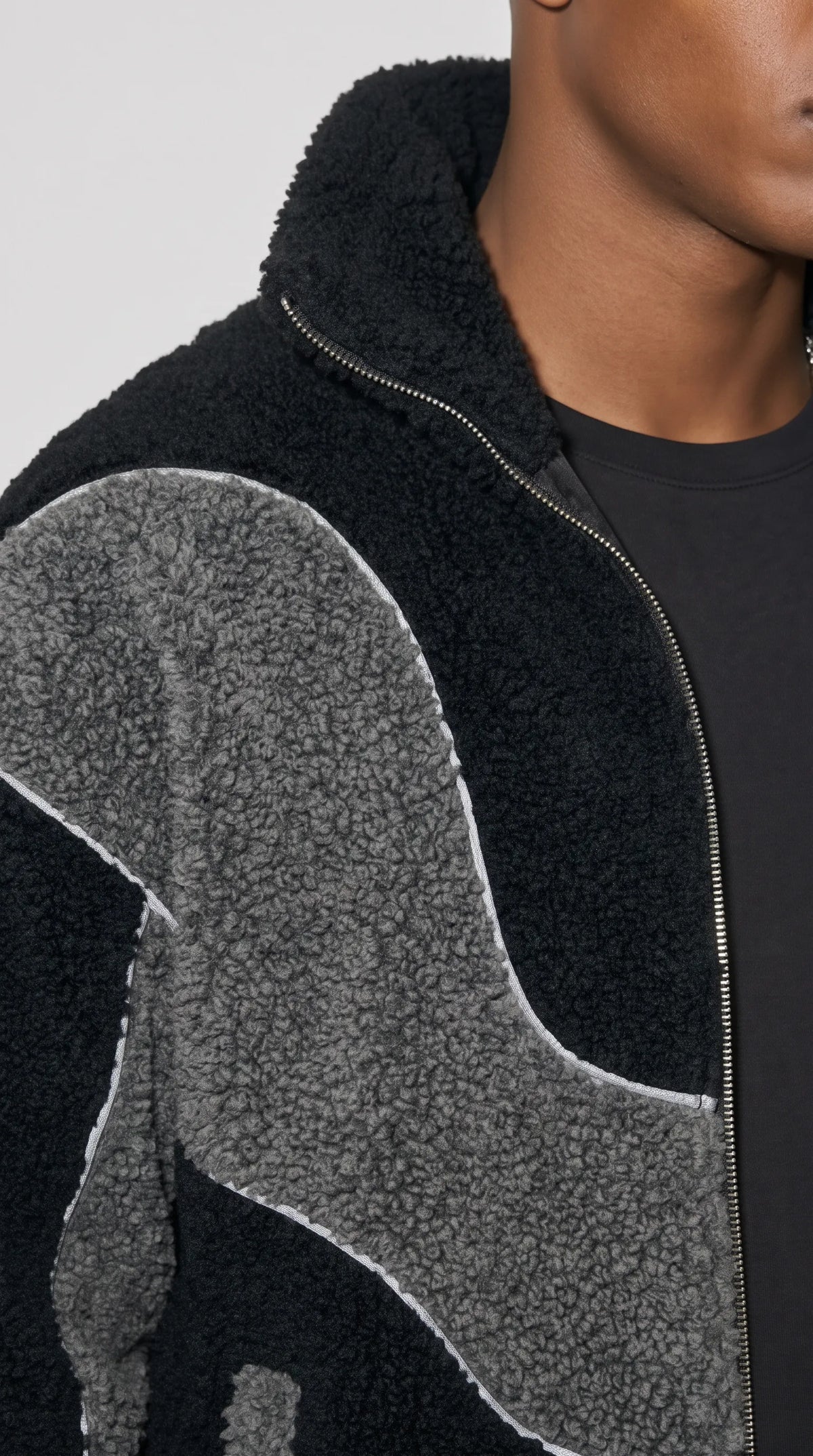 Detailed styling of a black and charcoal-grey wool-blend fleece jacket, highlighting its dense curly pile texture, smooth metal front zipper and crisp white piping around the flowing contrast panels. The interplay between tactile dark fleece and sculptural grey sections gives this designer-inspired outerwear a distinctive retro streetwear character with carefully defined construction details.