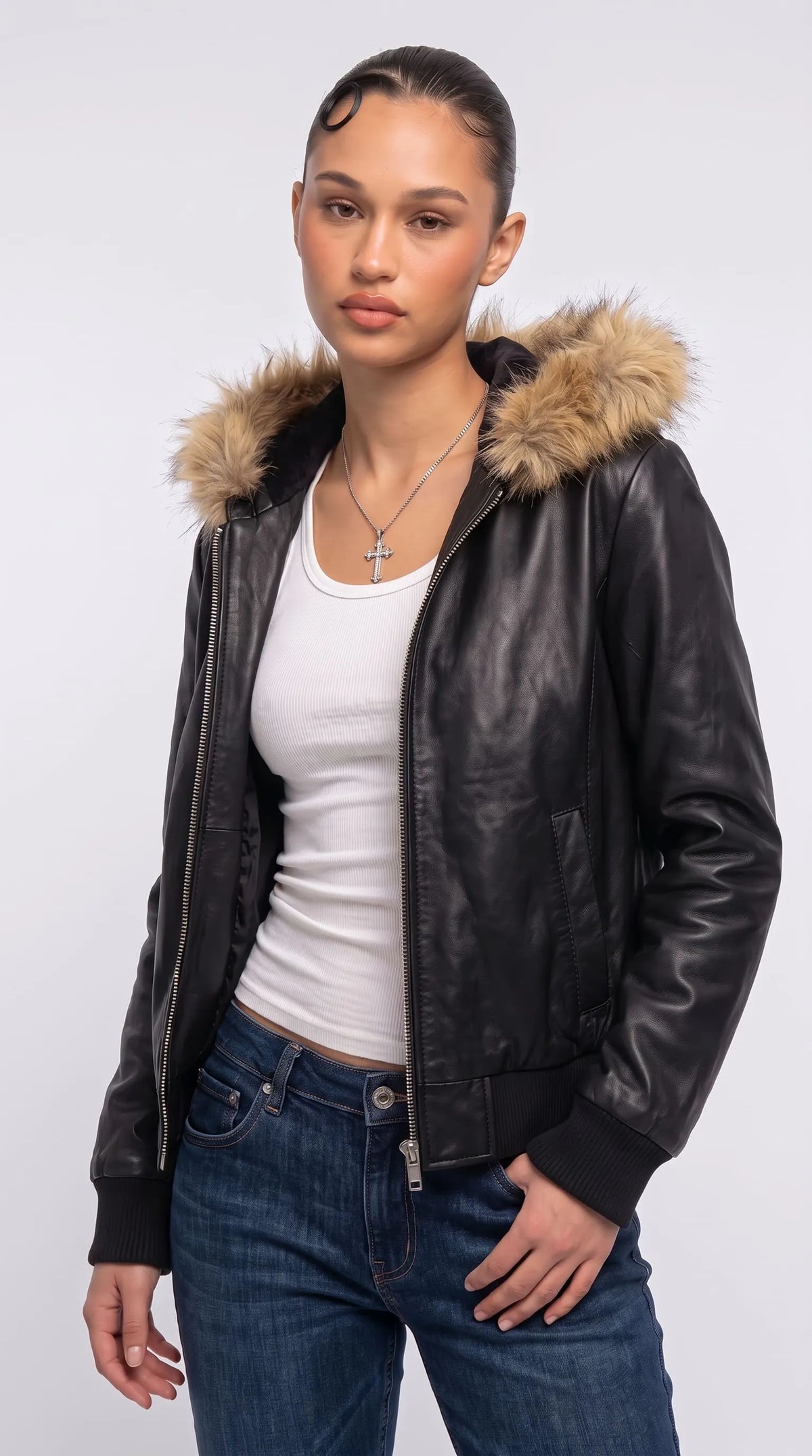 Oversized dark black genuine leather Y2K bomber jacket with a thick tan-brown faux-fur-trimmed hood, ribbed cuffs and a structured waistband, styled with a white ribbed tank top, blue denim jeans and a silver cross necklace. The relaxed dropped-shoulder fit and contrasting warm fur create a distinctive vintage streetwear look with a bold, modern winter-layering aesthetic.