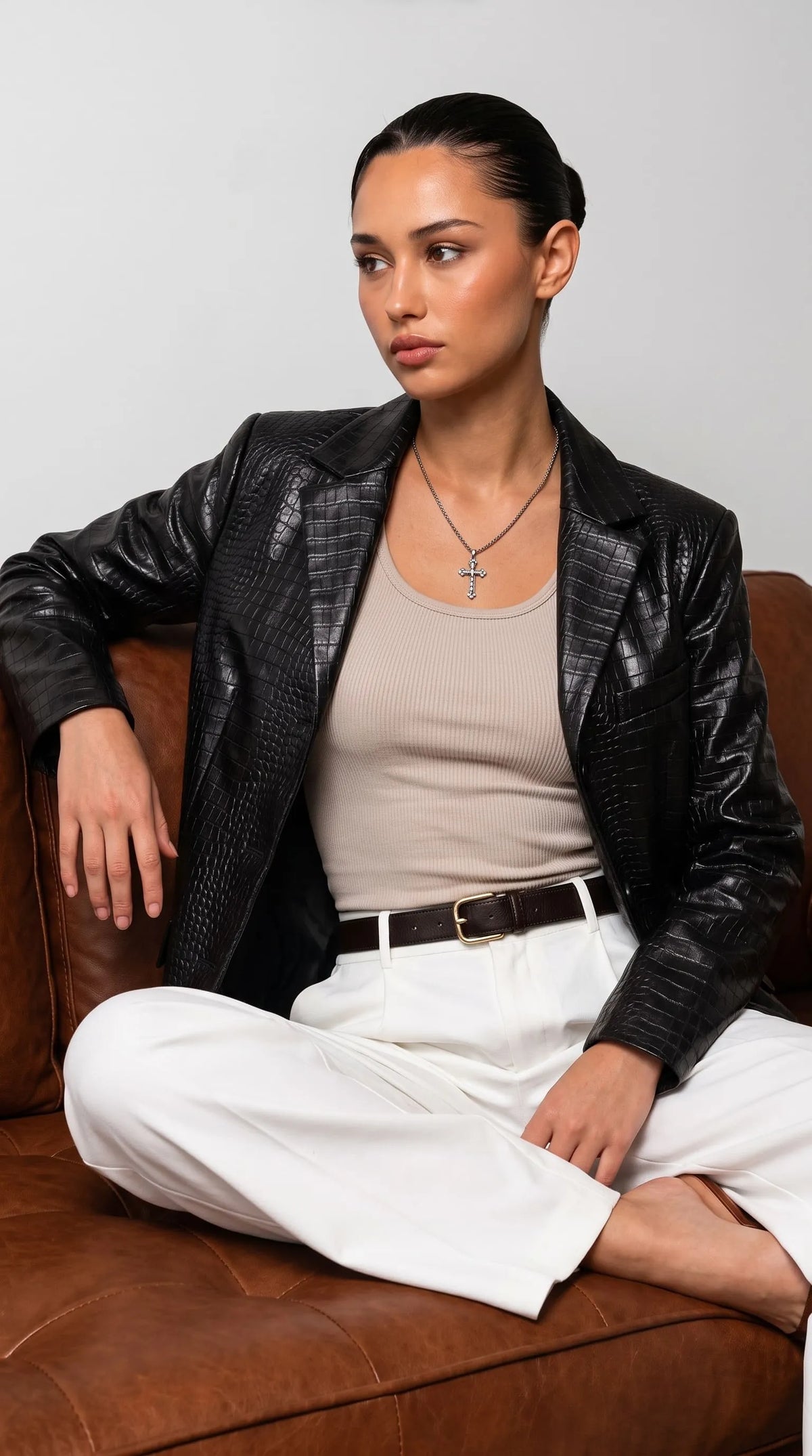 Editorial women's fashion featuring a glossy black crocodile-embossed leather blazer paired with a fitted neutral top, white trousers, a dark belt and delicate silver jewellery while seated on a cognac leather sofa. The bold textured finish contrasts with the clean tailored outfit, bringing a confident designer-inspired mood to contemporary luxury dressing, sophisticated street style and evening-ready looks.