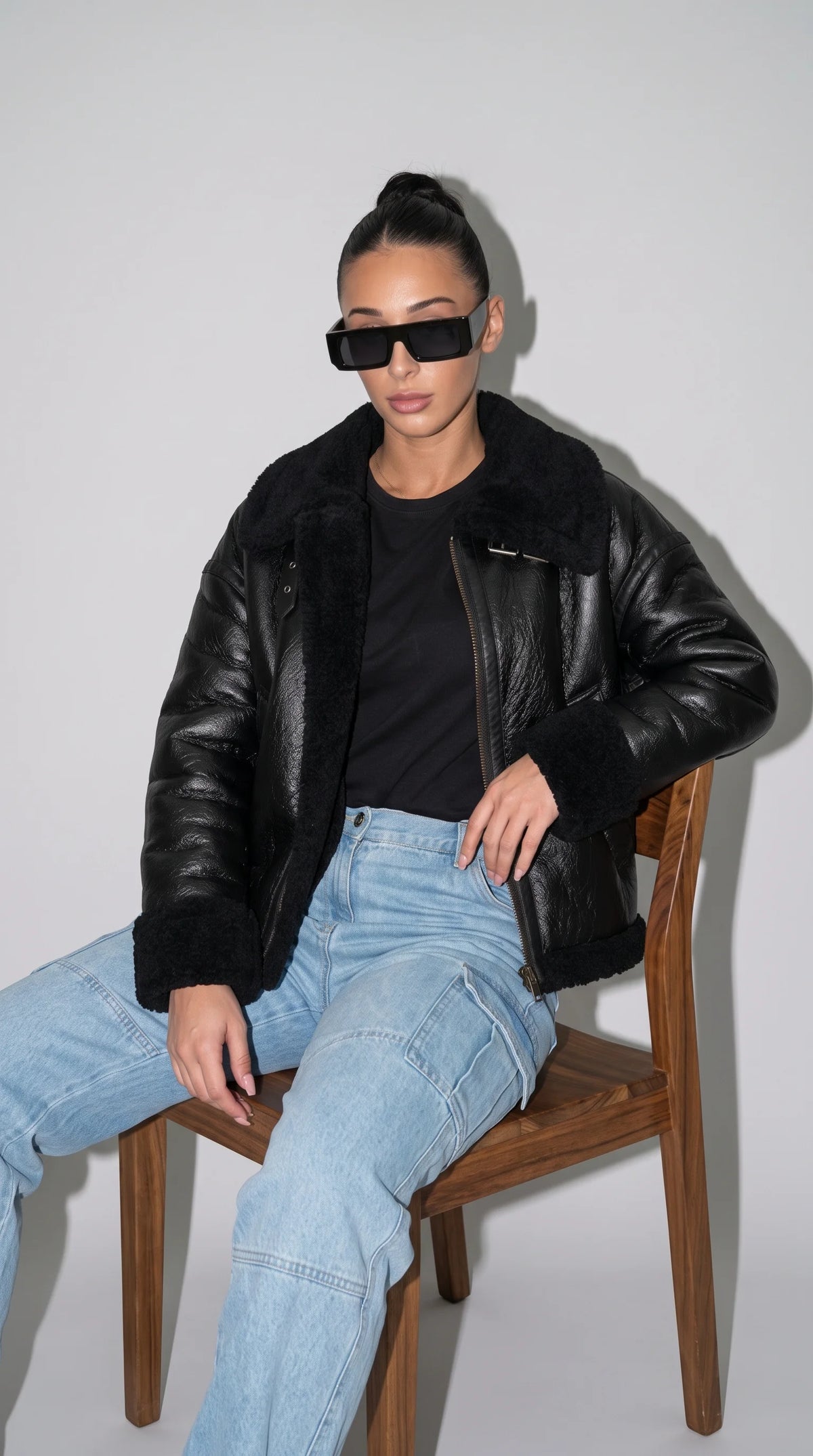 Black faux-shearling-lined leather flight jacket styled with a plain black top and light-blue cargo jeans, combining a plush aviator collar, thick textured cuffs, glossy leather panels and an asymmetric front closure for a relaxed utility-inspired streetwear outfit.