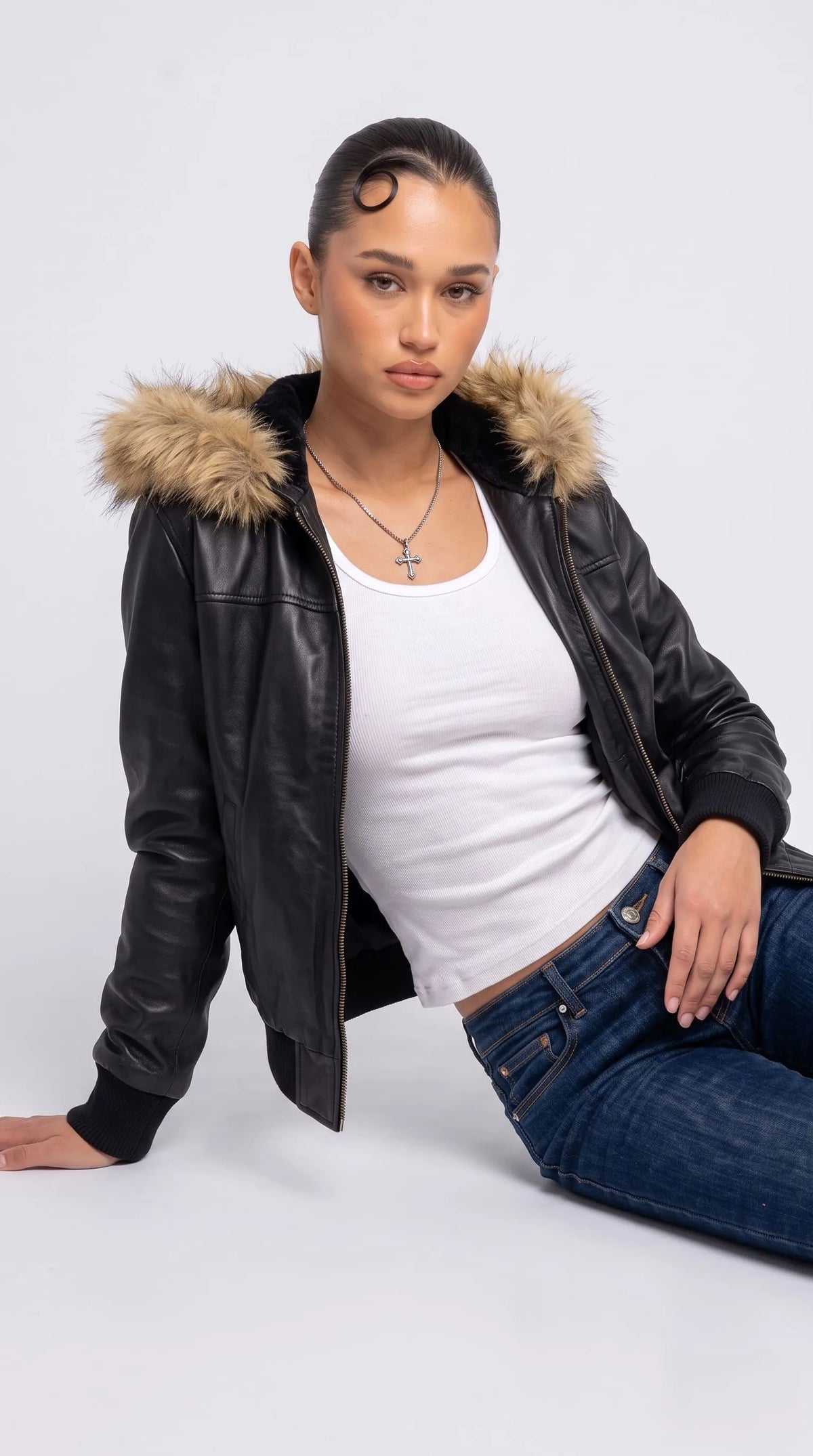 Fashion editorial featuring a black genuine leather oversized bomber jacket with a fluffy brown faux-fur hood, worn open over a white fitted ribbed top and dark-blue jeans while reclining in a relaxed studio pose. The voluminous sleeves, metallic front zipper and contrasting hood trim give this Y2K-inspired outfit a confident casual attitude, combining classic denim styling with statement leather outerwear.