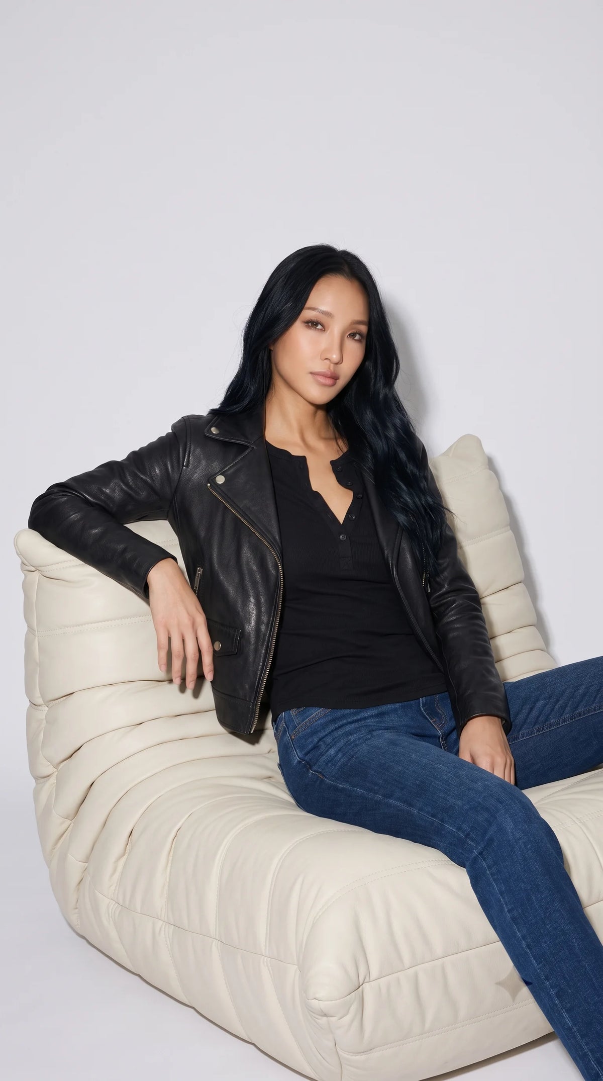 Black leather moto jacket styled with a fitted black ribbed shirt and classic blue denim in a relaxed studio editorial, showcasing its asymmetric front zip, broad lapels, silver snap accents and multiple practical pockets for elevated everyday fashion.