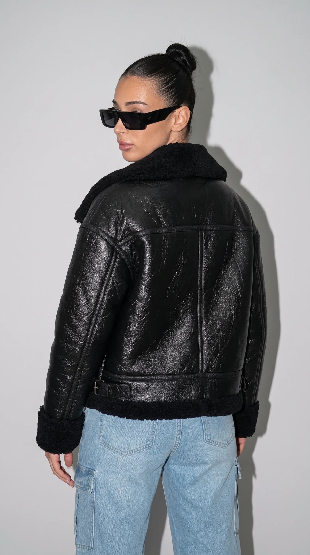 Black leather aviator jacket with a stitched shoulder yoke, defined centre-back panel, adjustable waist tabs and plush black faux-shearling trim along the hem, showcasing its structured construction and distinctive vintage flight-jacket character.