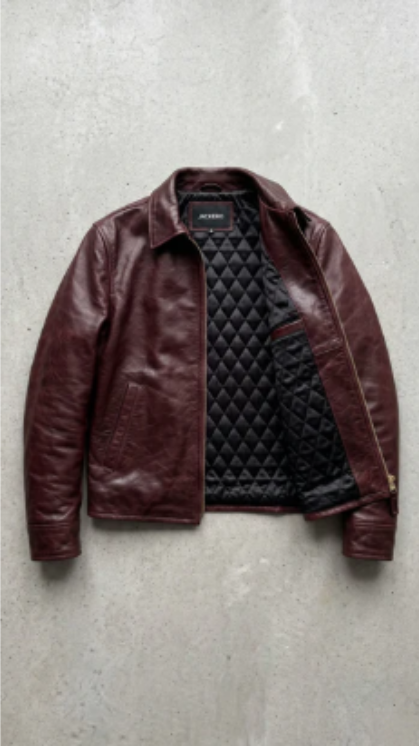 A burgundy leather jacket with a black diamond-quilted lining, laid flat on a textured gray concrete surface.