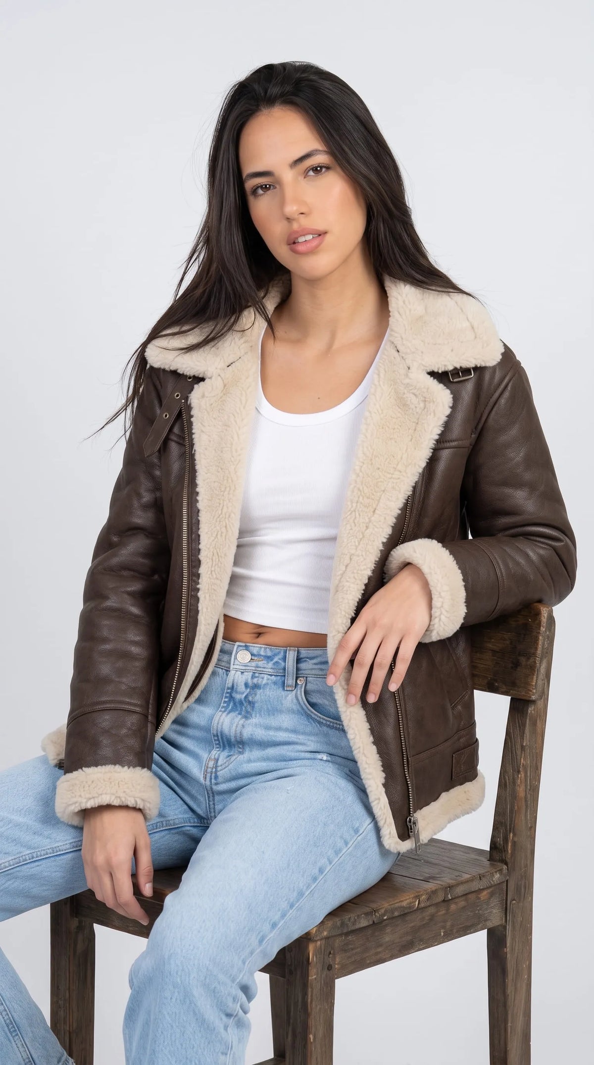 Contemporary outfit featuring a dark chocolate-brown leather aviator jacket with plush cream shearling lapels and cuffs, worn over a white crop top with light-blue jeans in relaxed seated styling. The oversized sleeves, open front zipper and contrasting fluffy trim create a confident editorial look that balances rugged outerwear with clean minimal basics for modern casual fashion.