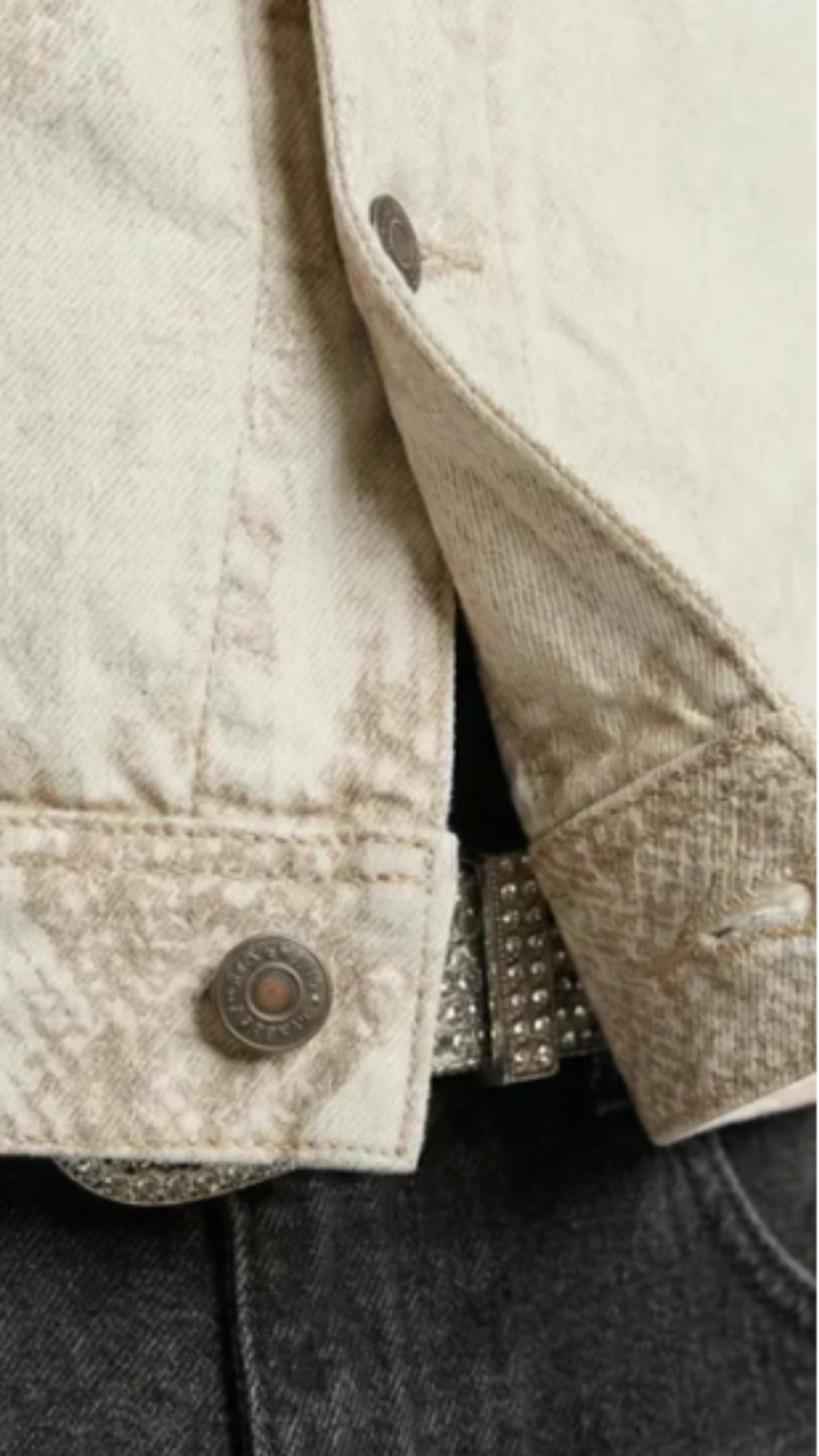 Close-up of a light beige textured denim jacket with a snakeskin-patterned cuff, paired with dark gray jeans and a studded belt.