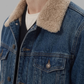 Close-up of a person wearing a blue denim jacket with a thick beige sherpa collar over a white t-shirt, against a solid gray background.