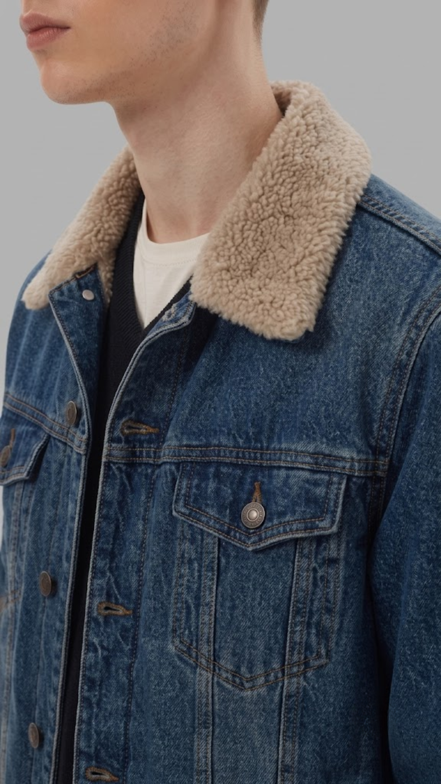 Close-up of a person wearing a blue denim jacket with a thick beige sherpa collar over a white t-shirt, against a solid gray background.