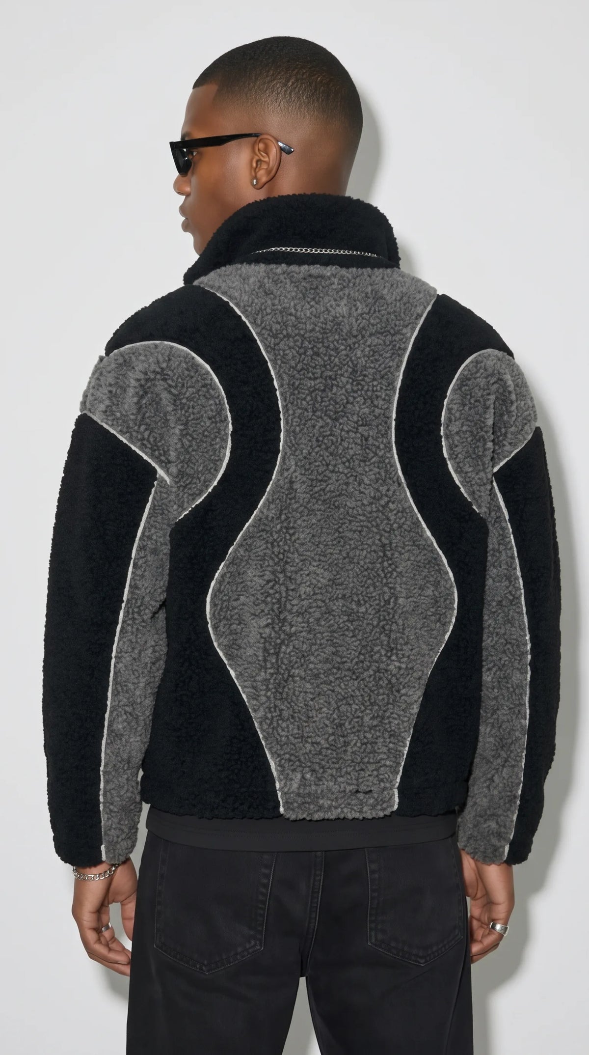 Black and charcoal-grey textured fleece jacket showcasing an eye-catching curved back panel, contrasting black sleeves and precise white piping along the sculpted seams. The oversized Y2K-inspired shape pairs a distinctive retro colour-block pattern with a relaxed urban silhouette, making it an expressive layering choice for modern grunge outfits and alternative street fashion.