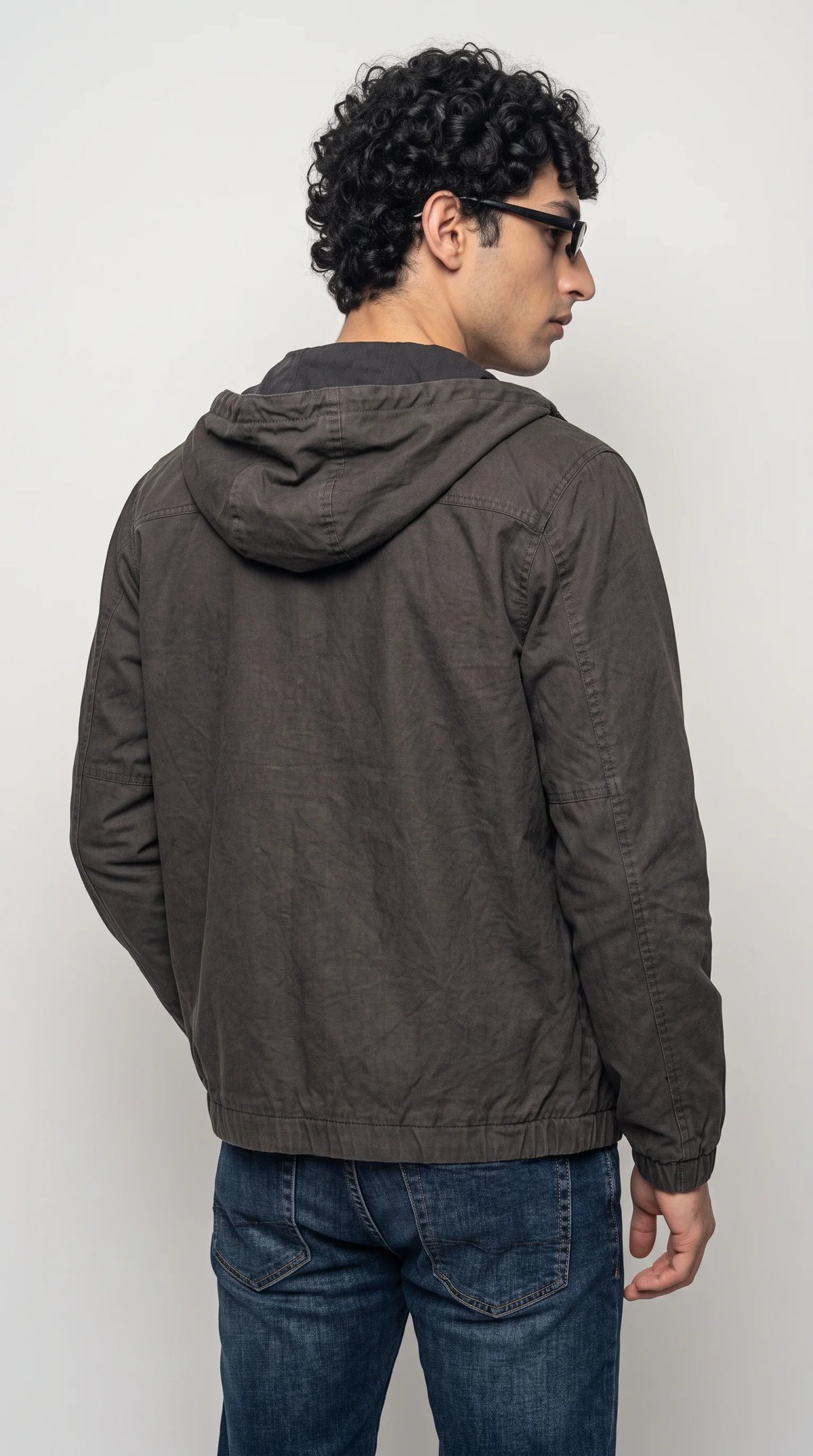 Dark-grey silk-cotton hooded jacket with a practical attached hood, structured shoulder seams, long sleeves and a gathered elasticated hem that creates a neat casual silhouette. Paired with dark denim, this understated utility-style outer layer brings a rugged modern feel to daily outfits while keeping the overall look clean, versatile and easy to coordinate.