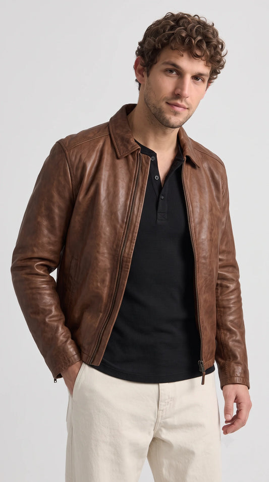 Men's chestnut-brown genuine leather jacket with a rich naturally textured finish, classic pointed collar and full-length metal zipper, styled open over a black ribbed henley shirt with ivory trousers for a polished smart-casual outfit.