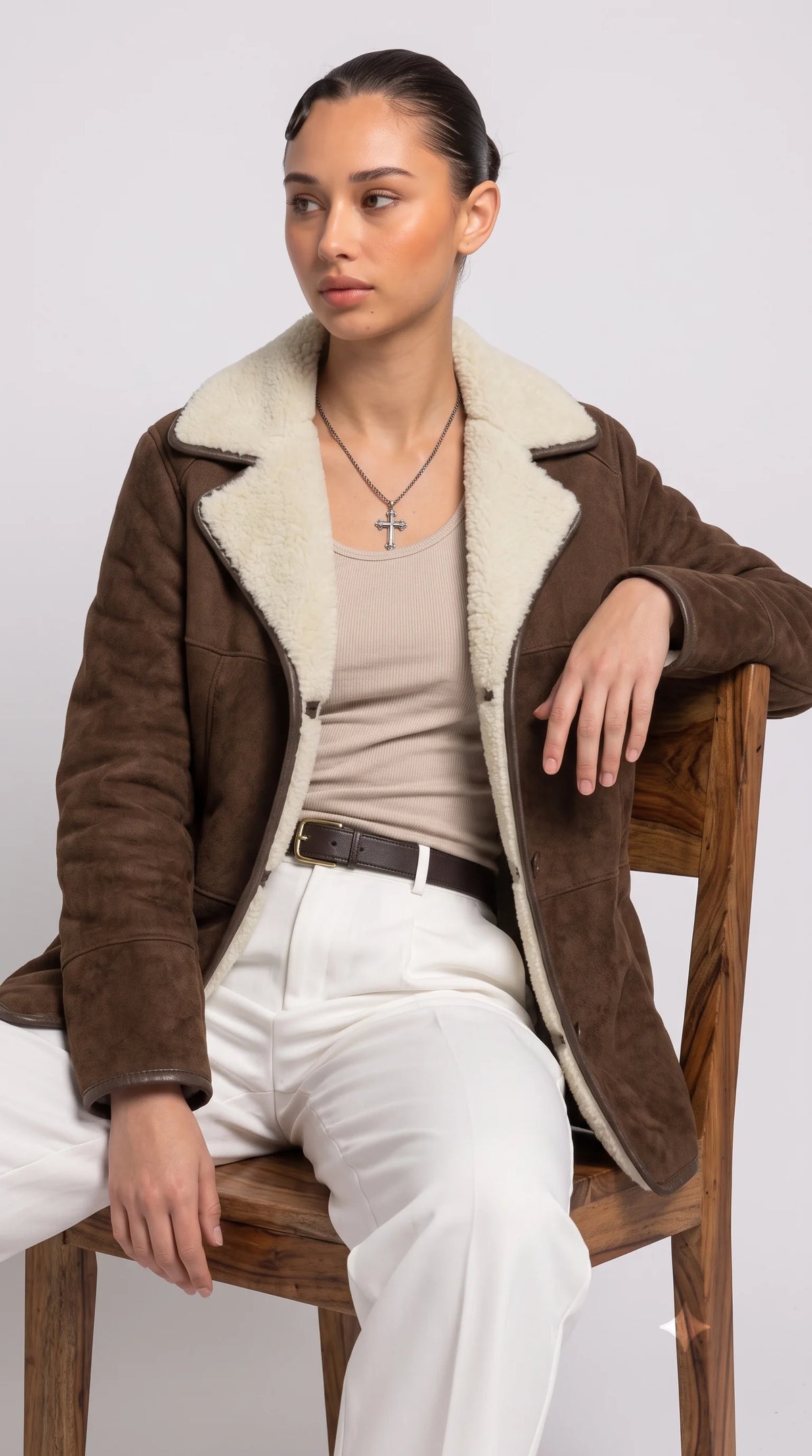 Dark chocolate-brown suede coat styled with a beige ribbed top and ivory tailored trousers in a relaxed luxury editorial, showcasing its oversized cream faux-shearling collar, buttoned front, contrasting dark piping and plush interior for an elegant neutral winter outfit.