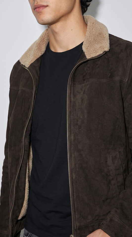 A dark brown suede shearling jacket with a plush cream collar is layered over a knitted sweater to create a relaxed, contemporary winter outfit. Its clean zip-front design, rich coffee-brown color and structured shoulders combine rugged outdoor character with refined city streetwear, making it an appealing choice for everyday cold-weather layering.