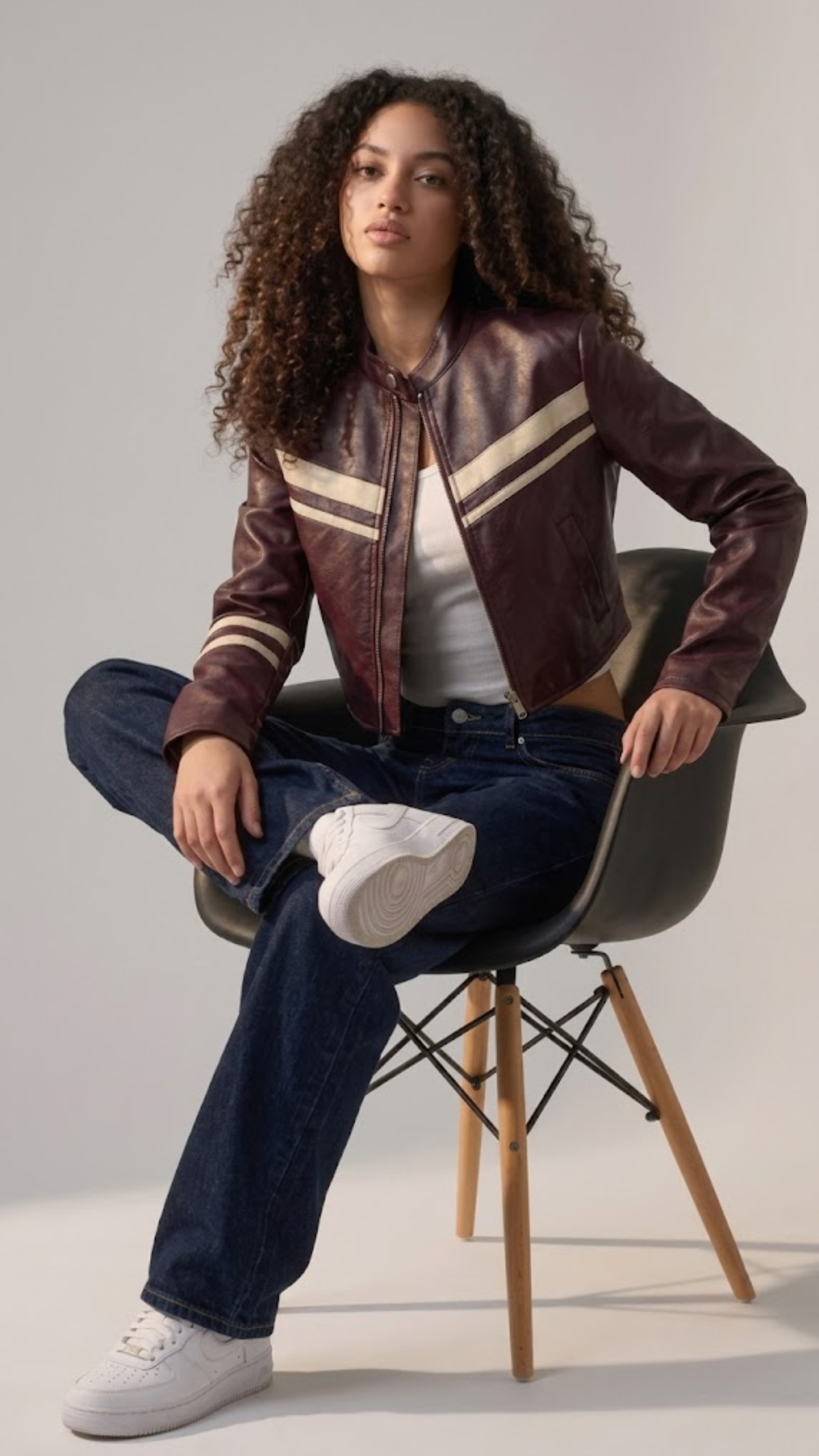 A man wearing a black leather jacket, white shirt, black tie, and dark pants sits on a wooden chair against a plain light gray studio background.