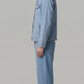 A man shown in profile wearing a light blue denim jacket and matching light blue denim jeans, paired with brown and white sneakers, against a solid light gray studio background.