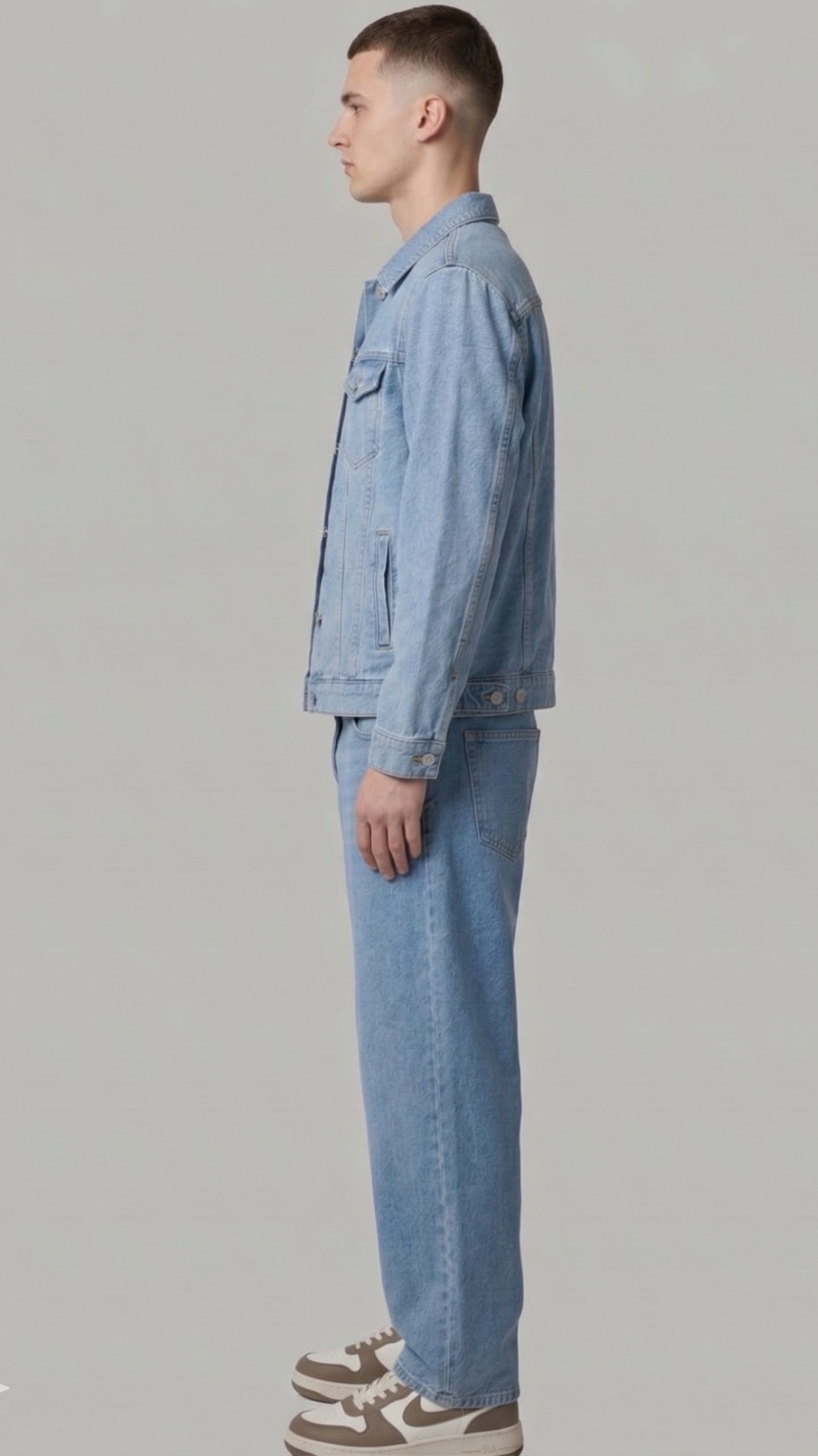 A man shown in profile wearing a light blue denim jacket and matching light blue denim jeans, paired with brown and white sneakers, against a solid light gray studio background.