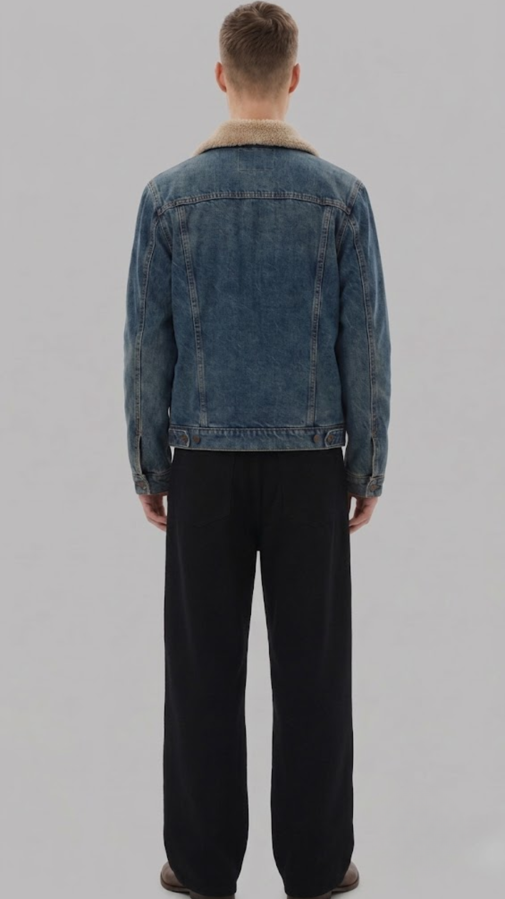 A man viewed from behind wearing a dark gray washed denim jacket with a white shearling collar, paired with dark gray jeans and brown shoes, against a light gray studio background.