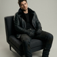 Man wearing a black leather jacket sitting on a black chair against a gray background