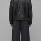 A man viewed from the back wearing a black leather jacket and loose black trousers, standing against a plain light gray studio background.