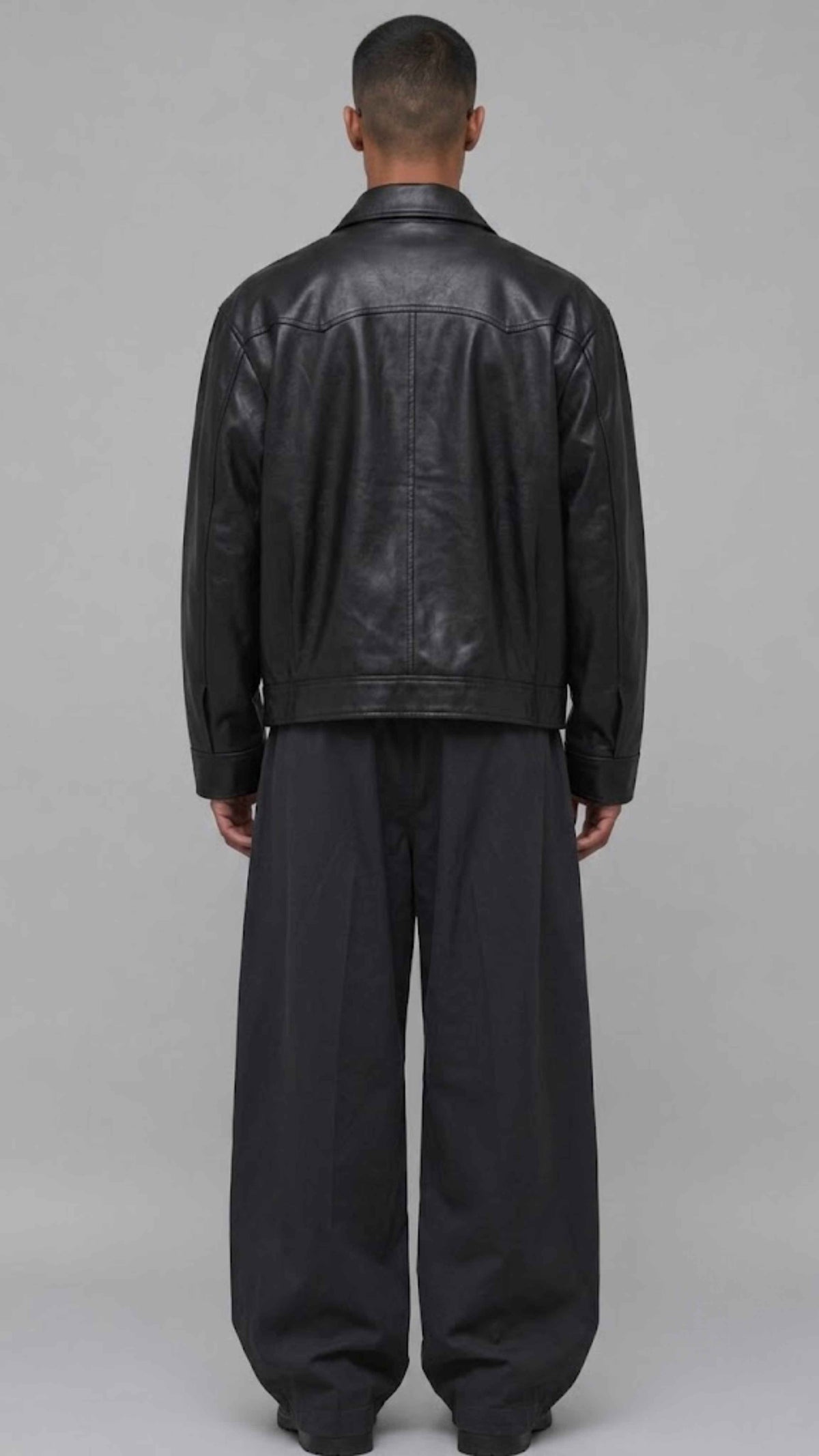 A man viewed from the back wearing a black leather jacket and loose black trousers, standing against a plain light gray studio background.
