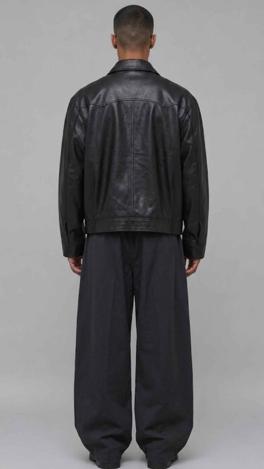 A man viewed from the back wearing a black leather jacket and loose black trousers, standing against a plain light gray studio background.