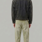 A man viewed from behind wearing a dark charcoal grey denim jacket and blue jeans, standing against a light gray studio background.