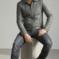 Man wearing a gray leather jacket and jeans sitting on a concrete block against a plain background