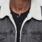 A close-up side view of a person wearing a dark washed denim jacket over a white shirt and light olive pants, against a plain gray studio background.