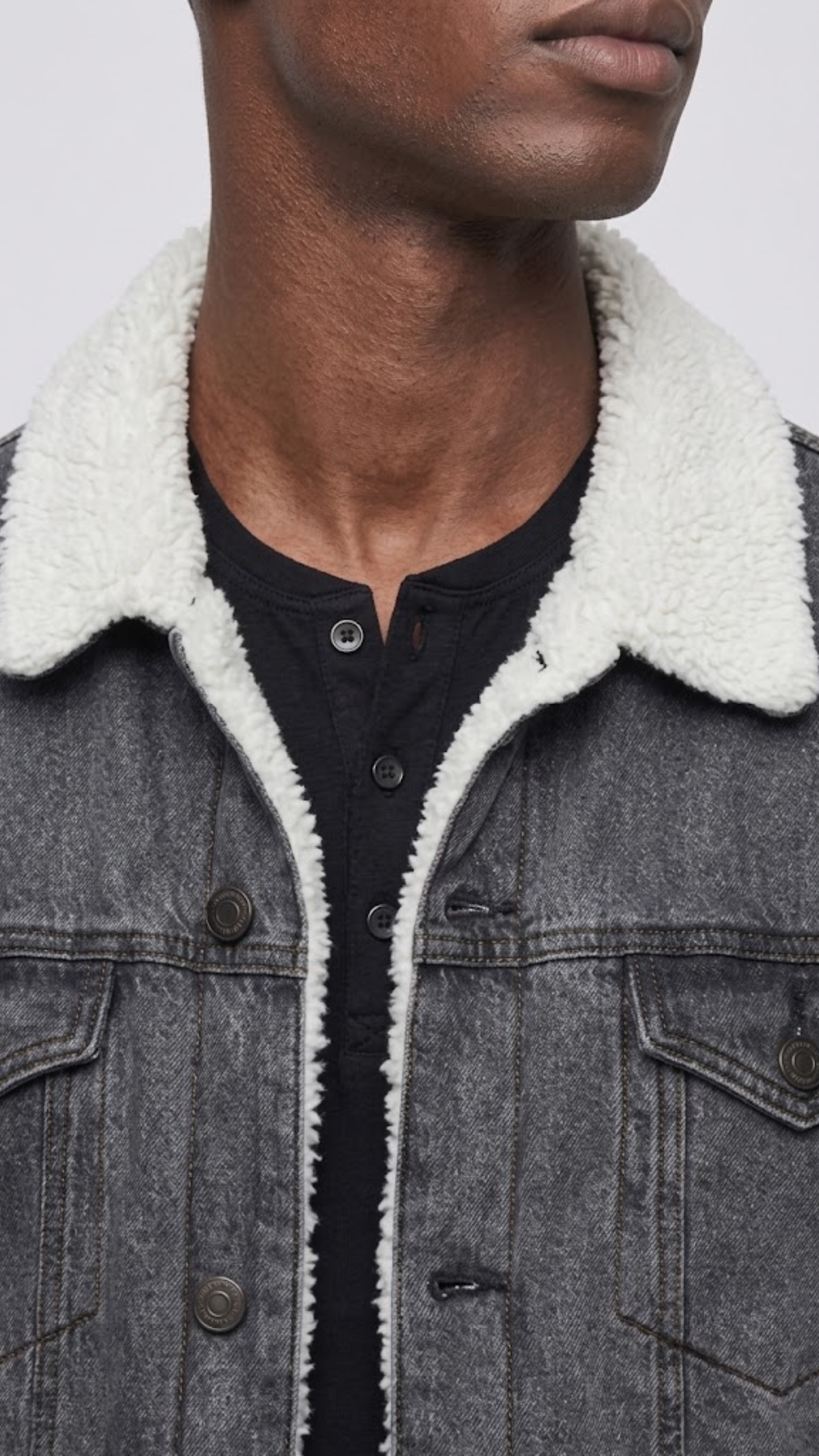 A close-up side view of a person wearing a dark washed denim jacket over a white shirt and light olive pants, against a plain gray studio background.