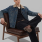 A man wearing a dark gray denim jacket with a white shearling-style collar, dark gray jeans, and brown suede boots, sitting on a wooden chair against a light gray studio background.