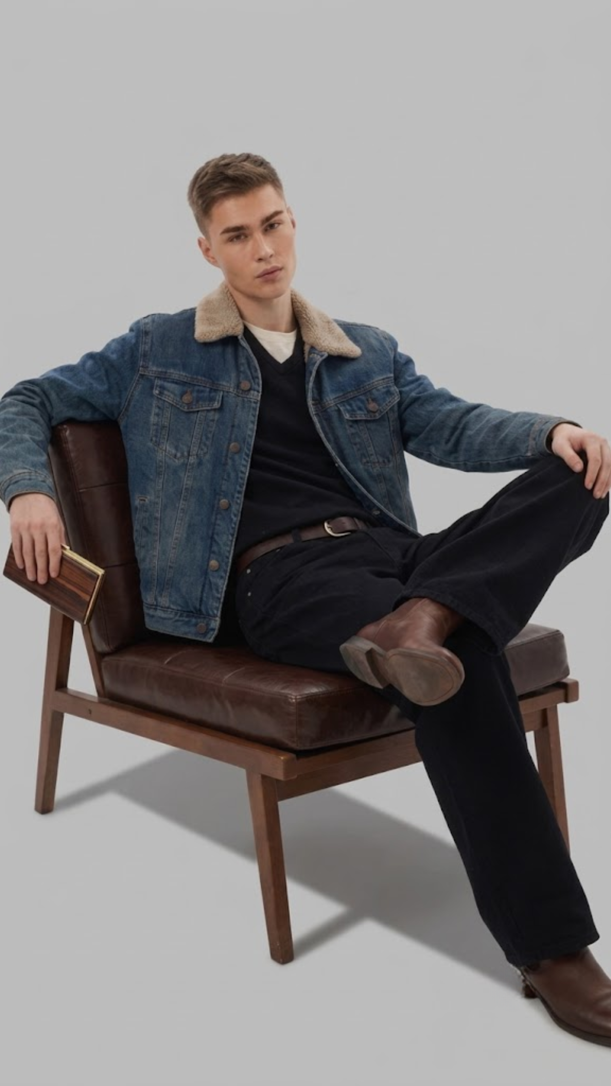 A man wearing a dark gray denim jacket with a white shearling-style collar, dark gray jeans, and brown suede boots, sitting on a wooden chair against a light gray studio background.
