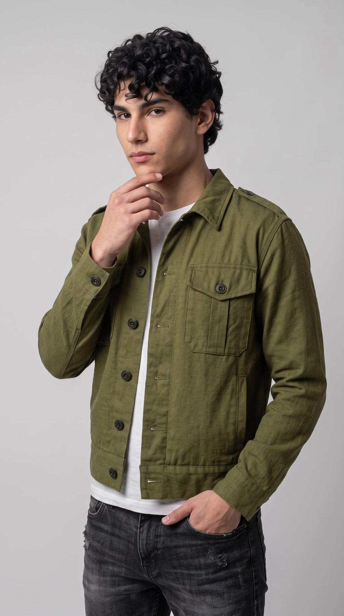 Cropped olive green cotton canvas jacket with a tailored waist-length cut, military shoulder straps, a single-breasted black button closure and structured chest pockets, paired with washed black jeans for a contemporary utilitarian streetwear outfit.