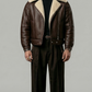 a model wearing a premium dark brown leather aviator jacket with a thick cream shearling collar, standing against a clean grey studio backdrop.
