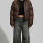 a model wearing a premium dark chocolate brown puffer jacket with clean horizontal quilting, standing against a solid grey studio background.