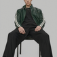 Man sitting on a chair wearing a green jacket with white stripes and black pants on a gray background